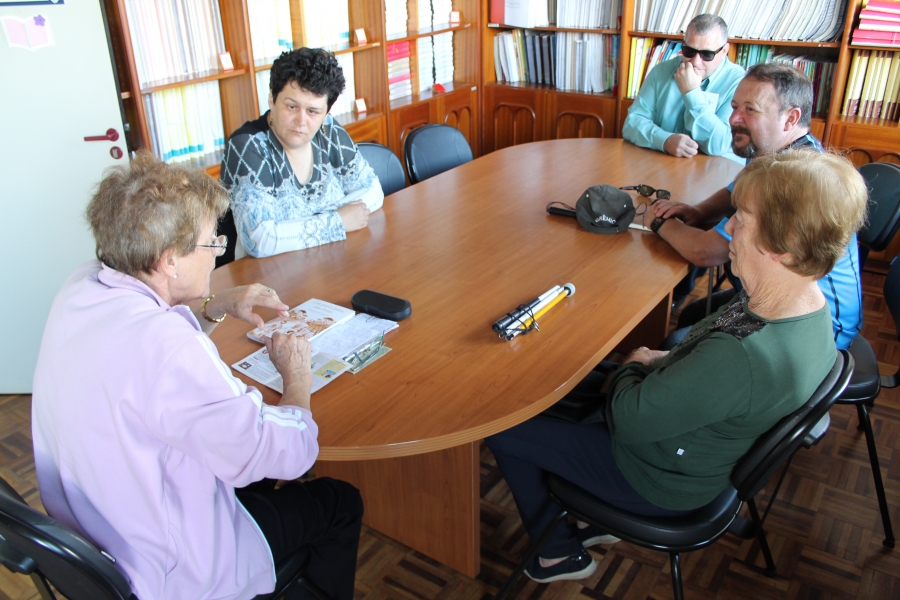 Imagem: Grupo de usuários adultos sentados em torno da uma mesa central. Na ponta desta mesa, profissional com alguns livros a sua frente para iniciar as leituras aos presentes.