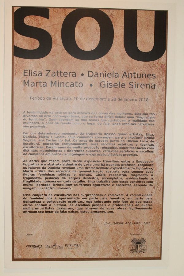 Imagem: Placa no formato retangular, com inscrições do nome da exposição, das artistas responsáveis pelas peças e uma breve descrição sobre o propósito da exposição.