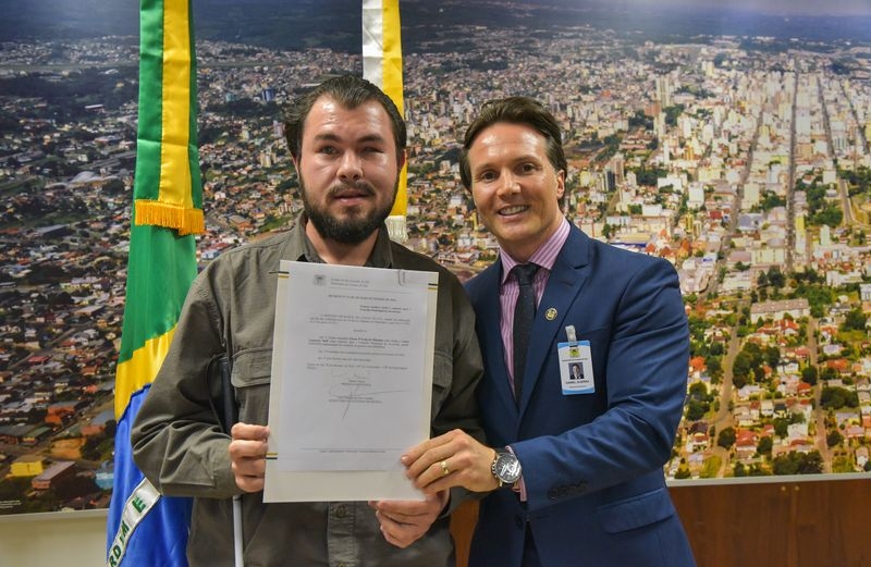 Imagem: Édson está recebendo das mãos do prefeito Daniel Guerra, o documento de posse. Ao fundo hasteada a bandeira nacional e a bandeira municipal. Atrás das bandeiras uma foto grande da cidade de Caxias do Sul.